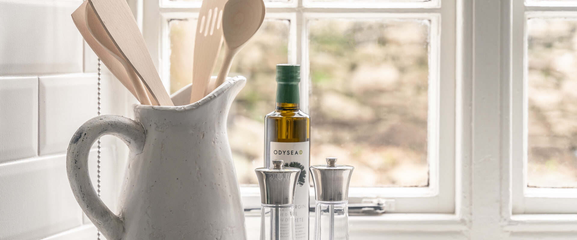 Close up of a kitchen counter with a white jug with wooden cooking spoons inside. On the right are salt and pepper mills, and a bottle of pomegranate molasses.